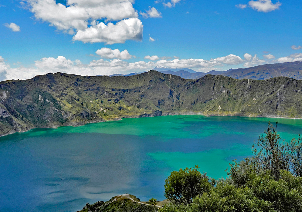 Quilotoa Lake Ecuador View of Quilotoa Lake’s vibrant waters framed by dense greenery and distant volcano peaks under puffy clouds