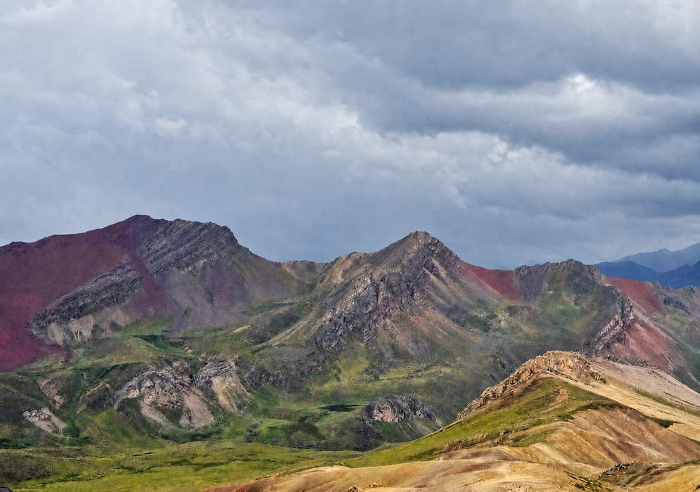 Jagged Andean peaks colored with stripes of red, purple, and olive green seen beneath an ominous stormy sky in the Rainbow Mountain range
