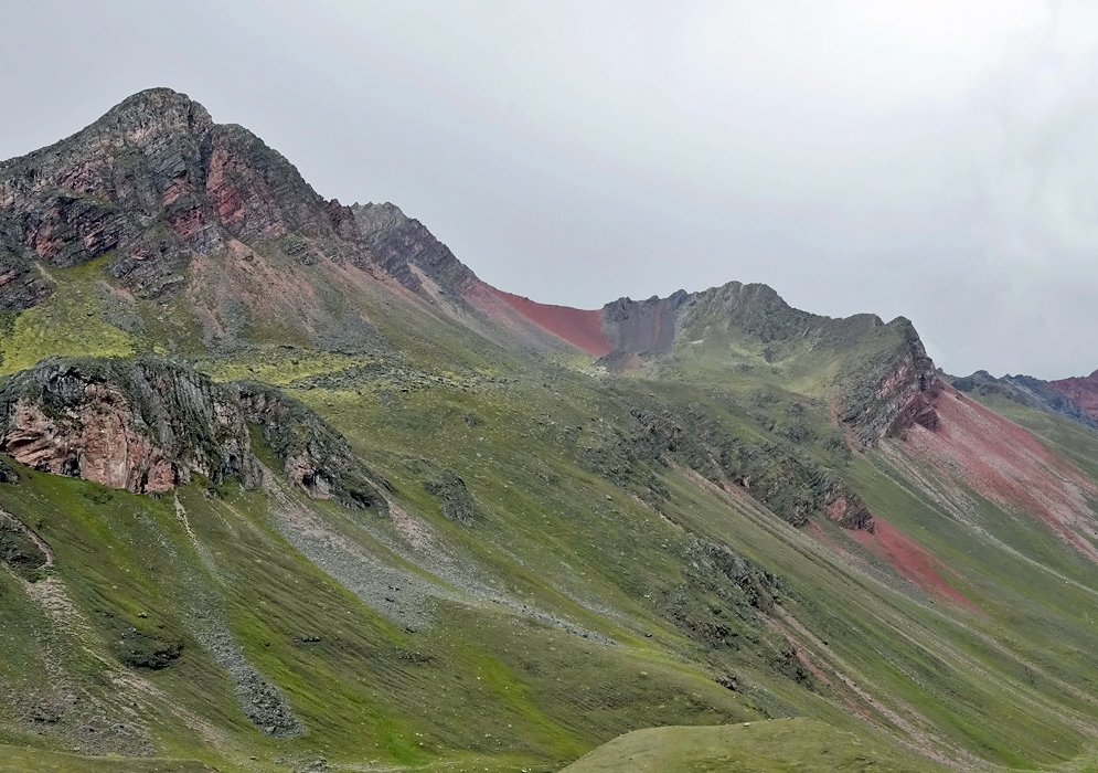 A rugged green mountainside in the Andes of Peru, streaked with red and pink mineral layers rising sharply under a cloudy sky