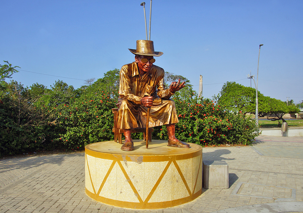 A golden statue of a seated man with a hat, cane and glasses under blue skies