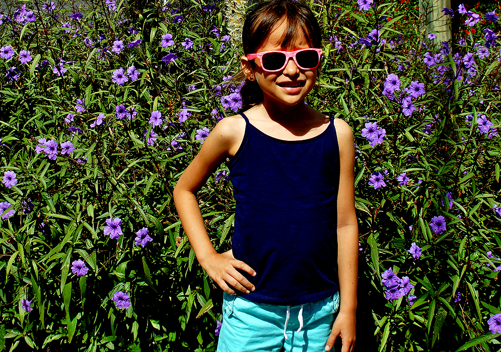 Smiling girl with sunglasses standing in front of a Ruellia brittoniana flowers in sunlight