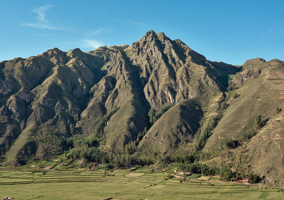 Wide landscape of the Sacred Valley of the Incas, showing layered mountains, a riverbed, farmlands, and a misty sky over the Andes