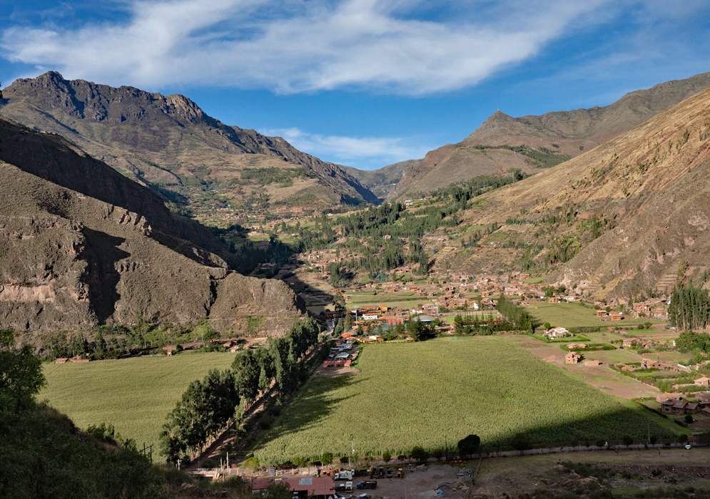 Wide landscape of the Sacred Valley of the Incas, showing layered mountains, a riverbed, farmlands, and a misty sky over the Andes
