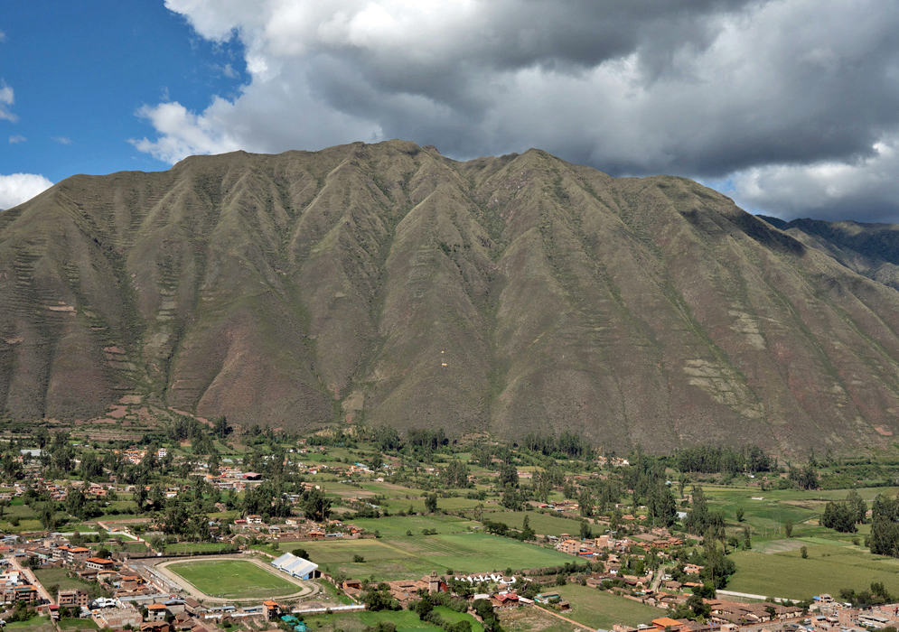 Panoramic high-angle shot of the Urubamba area in the Sacred Valley, with steep green slopes, scattered villages, and cloud patches over the Andes.