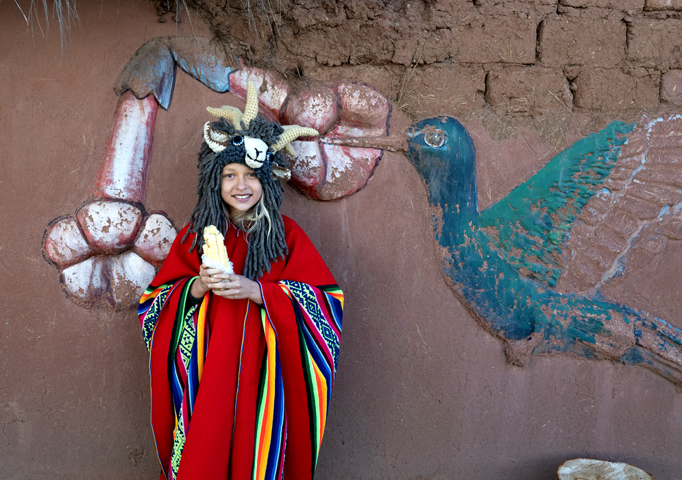 Girl in Traditional Dress at Sacsayhuamán, Cusco. Girl in a red Andean poncho and animal hat holding white corn in front of a colorful wall mural at Sacsayhuamán near Cusco, Peru