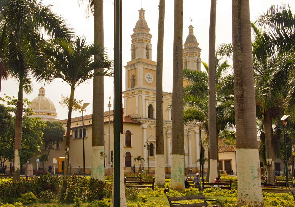A tan-color church with two towers behind a park with palm trees