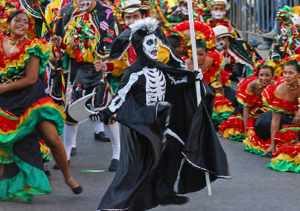 Barranquilla Carnival Performer in Skeleton Costume Person dressed as a skeleton with black and white face paint and a dramatic costume, marching with a staff during the Barranquilla Carnival parade, surrounded by colorful dancer