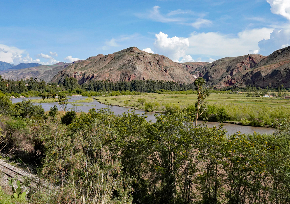 Wide view of the Vilcanota River winding through a green rural valley in the Andes, with forested banks, small farms, and red-tinted mountains in the background