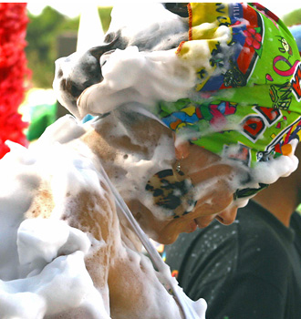 Woman wearing a colorful Carnival hat looks downward as thick white foam covers her face, hair, neck, and shoulders